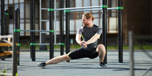 Slanke man in sport outfit voert een stretch oefening uit om zijn adductoren te rekken. Dit doet hij buiten op een trainingsveld